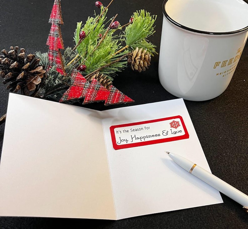 Peut inclure: Une carte de v&oelig;ux blanche ouverte avec une &eacute;tiquette rouge indiquant "It's the Season for Joy, Happiness & Love". Un stylo blanc avec des d&eacute;tails dor&eacute;s repose sur la carte. Une tasse blanche et des d&eacute;corations de No&euml;l sont en arri&egrave;re-plan.