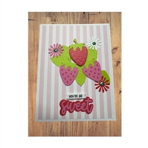 May include: A handmade greeting card with a pink and white striped background. The card features three strawberries, green leaves, and decorative flowers. The text "YOU'RE SO Sweet" is displayed in red.