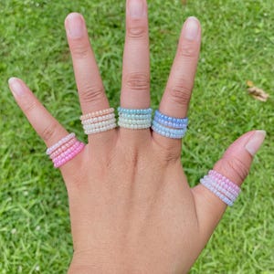 May include: A hand displays six beaded rings in shades of pink, peach, light blue, and lavender. The rings are made of small, colorful beads and are worn on multiple fingers. The background is a blurred green lawn.