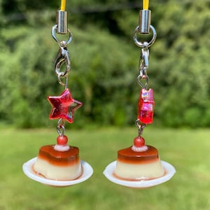 May include: Two food-themed phone charms. Each charm features a miniature dessert on a white plate, topped with a red bead. One charm has a red star, the other a red cube. Both are attached to a yellow cord.