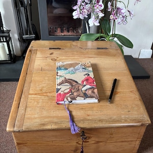 May include: A wooden chest with a hinged lid. A 2023 journal with a horse-riding scene and a black pen rest on top. A purple tassel hangs from the chest's lock. A fireplace and orchid plant are in the background.