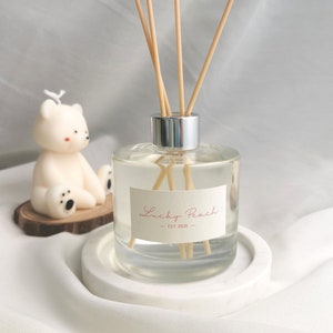 May include: A clear glass diffuser bottle with wooden reeds, a silver cap, and a label that reads "Lucky Peach - EST 2021." A white bear-shaped candle sits on a wooden slice. The diffuser sits on a white, round tray.