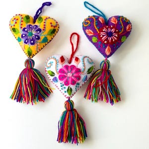 May include: Three heart-shaped ornaments with colorful embroidery and tassels. One is yellow with purple and green floral designs, another is purple with orange and red accents, and the third is white with pink flowers.