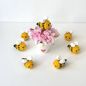 May include: Seven crocheted bumblebees in yellow and black with white wings, arranged around a small white pot with pink flowers.