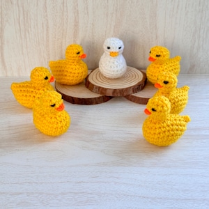 May include: A group of crocheted yellow duck figurines surrounding a single white crocheted duck figurine on a wooden base.