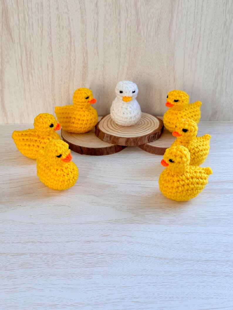 May include: A group of crocheted yellow duck figurines surrounding a single white crocheted duck figurine on a wooden base.