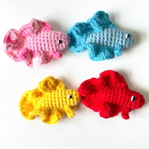 May include: Four crocheted fish appliques in pink, blue, yellow, and red. Each fish features a textured yarn body, wide flowing fins, and a small black and white bead eye. The set includes one of each color, arranged in a two-by-two grid pattern.