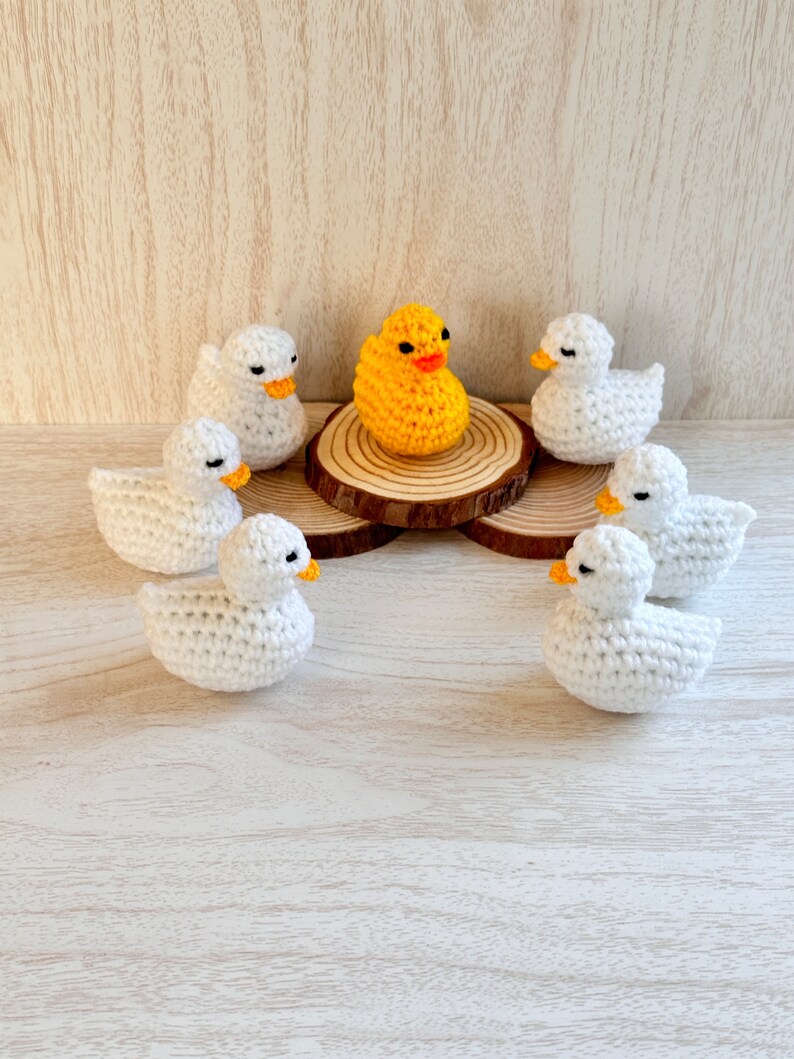 May include: A yellow crocheted duck surrounded by six white crocheted ducks on a wooden surface.