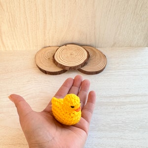 May include: A yellow crocheted duck toy held in a hand. The duck has black eyes and an orange beak.