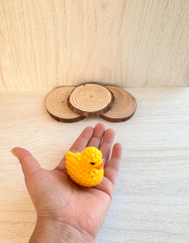 May include: A yellow crocheted duck toy held in a hand. The duck has black eyes and an orange beak.
