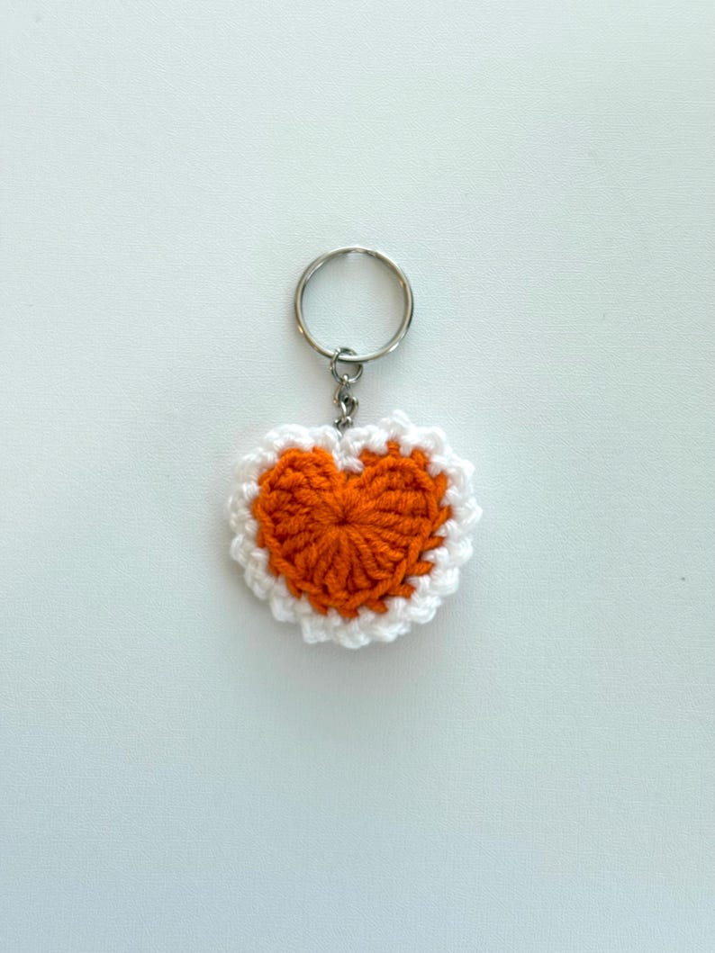 K&ouml;nnte beinhalten: Ein geh&auml;kelter Herz-Schl&uuml;sselanh&auml;nger. Das Herz ist orange mit einem wei&szlig;en Rand. Der Schl&uuml;sselanh&auml;nger hat einen silbernen Metallring zum Befestigen an Schl&uuml;sseln oder Taschen. Durchmesser ca. 7 cm.