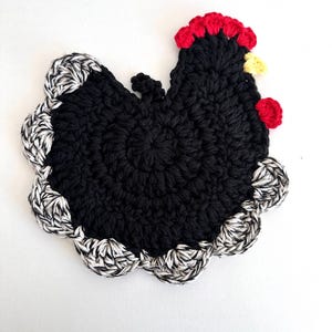 May include: A handmade, black crochet chicken-shaped trivet with a scalloped edge. The chicken has a red comb and wattle, and a yellow beak. The edge is a mix of black and white yarn. This trivet is designed to protect surfaces from hot items.