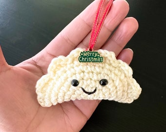 May include: A cream-colored crochet dumpling ornament featuring black safety eyes and a stitched smile. A small green metal charm with the text Merry Christmas is attached to the top, suspended by a thin red ribbon loop for hanging.