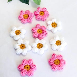 May include: A collection of handmade crochet flowers in pink and white, each with a yellow center. The flowers are arranged on a white surface, with green leaves in the background. These decorative flowers are ideal for crafting projects.