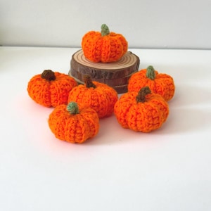 May include: A collection of six handmade crochet pumpkins in varying sizes. The pumpkins are a vibrant orange with green or brown stems. One pumpkin rests on a small wooden slice, while the others are arranged on a white surface. Perfect for fall decor.