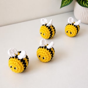 May include: Four small, round, crocheted bees featuring bright yellow bodies with horizontal black stripes, white wings, and small black eyes. Each bee is crafted with a textured yarn finish, creating a soft, spherical shape that sits upright.