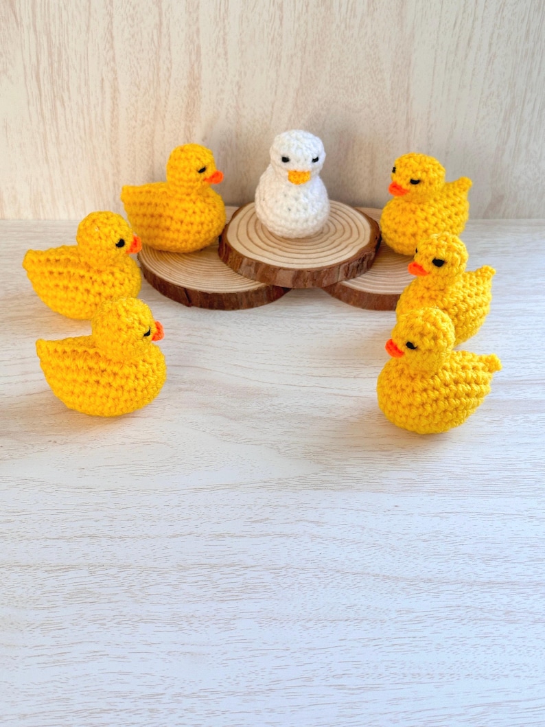 May include: A white crocheted duck surrounded by seven yellow crocheted ducks. The ducks are arranged in a circle around the white duck, which is sitting on a wooden log.