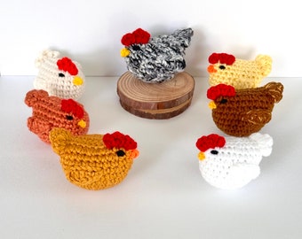 May include: A collection of seven crocheted chicken figurines in various colors, including white, yellow, brown, and gray. Each chicken has a red comb and beak. One chicken is displayed on a small wooden slice.