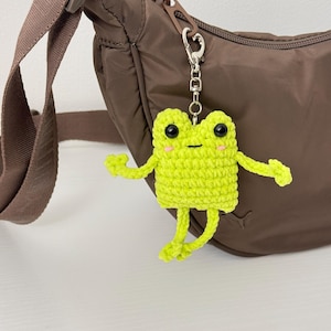 May include: A lime green crochet frog keychain with black eyes and pink cheeks. The frog has outstretched arms and legs. It is attached to a silver-colored metal clasp and is hanging in front of a brown purse.