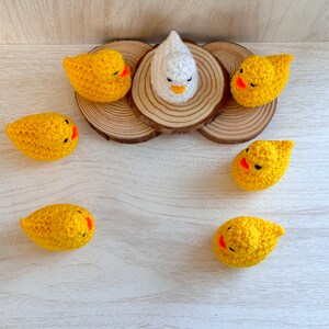 May include: A white crocheted chick surrounded by six yellow crocheted chicks on a wooden surface.