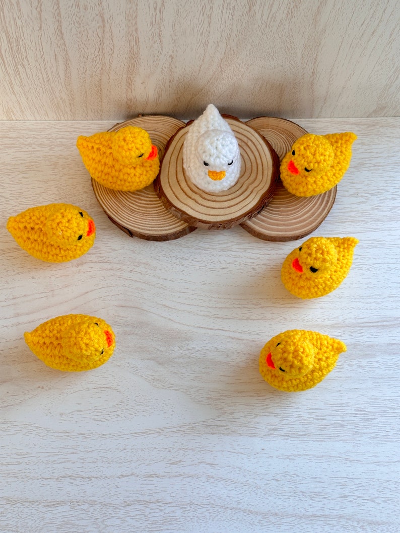 May include: A white crocheted chick surrounded by six yellow crocheted chicks on a wooden surface.