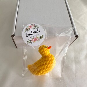 May include: A small yellow crocheted duck with an orange beak, packaged in a clear plastic bag with a white box and a sticker that says "handmade with love".