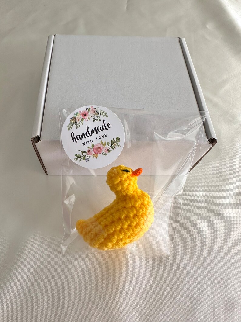 May include: A small yellow crocheted duck with an orange beak, packaged in a clear plastic bag with a white box and a sticker that says "handmade with love".
