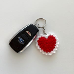 K&ouml;nnte beinhalten: Ein schwarzer Autoschl&uuml;ssel neben einem handgefertigten, rot-wei&szlig;en H&auml;kel-Herz-Schl&uuml;sselanh&auml;nger. Das Herz ist leuchtend rot mit einem wei&szlig;en, gewellten Rand, befestigt an einem silbernen Schl&uuml;sselring und einer Kette. Der Schl&uuml;ssel hat ein Subaru-Logo.