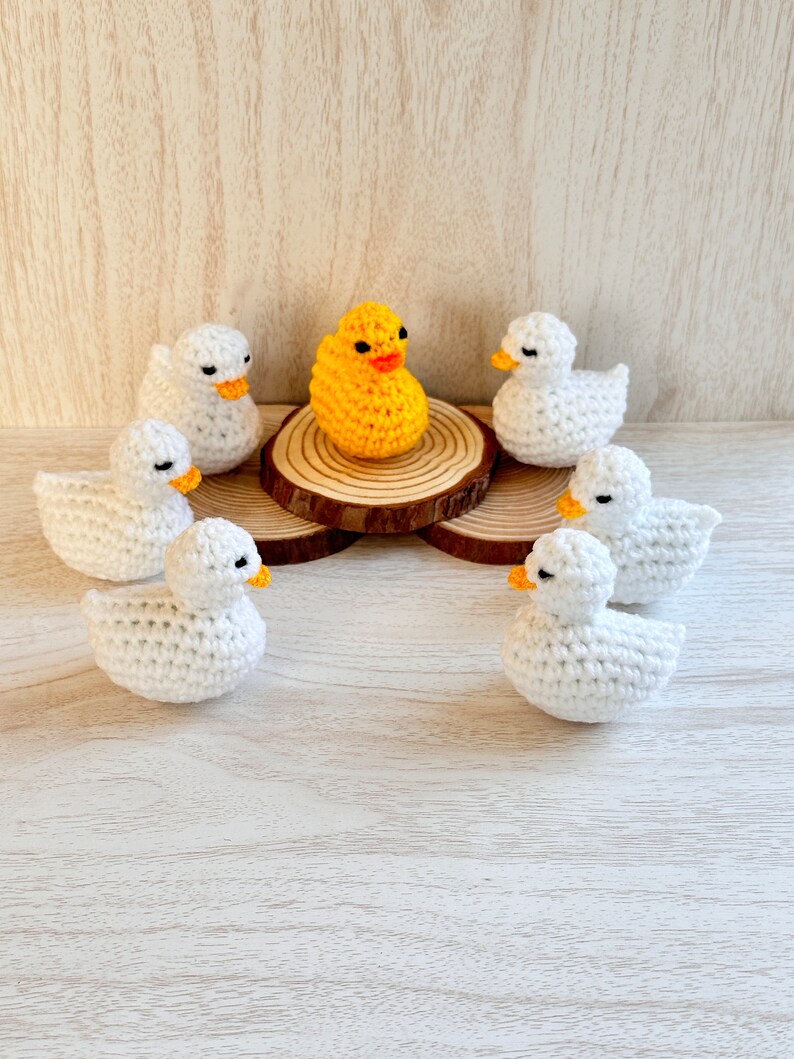 May include: A yellow crocheted duck surrounded by six white crocheted ducks on a wooden surface.