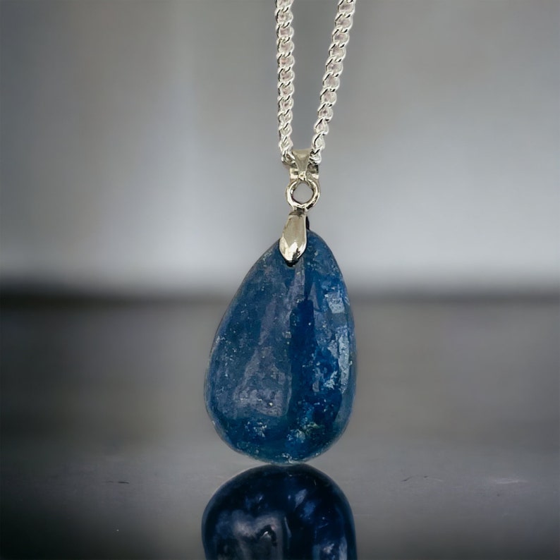 K&ouml;nnte beinhalten: Halskette mit silberner Kette und einem tropfenf&ouml;rmigen, tiefblauen Anh&auml;nger. Der Anh&auml;nger hat eine glatte, polierte Oberfl&auml;che und ist an einer silbernen &Ouml;se befestigt. Die Halskette wird vor einem reflektierenden Hintergrund gezeigt.