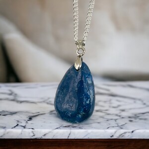 K&ouml;nnte beinhalten: Halskette mit blauem Edelstein-Anh&auml;nger und Silberkette. Der tropfenf&ouml;rmige Stein hat eine polierte, gl&auml;nzende Oberfl&auml;che. Die Halskette wird auf einer Marmoroberfl&auml;che pr&auml;sentiert.