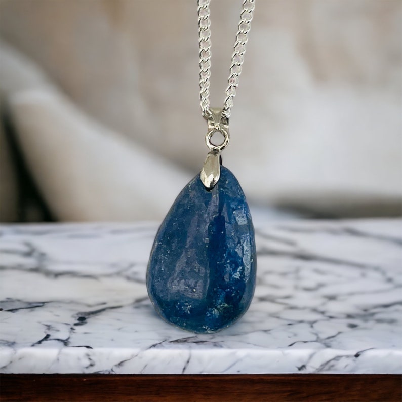 K&ouml;nnte beinhalten: Halskette mit blauem Edelstein-Anh&auml;nger und Silberkette. Der tropfenf&ouml;rmige Stein hat eine polierte, gl&auml;nzende Oberfl&auml;che. Die Halskette wird auf einer Marmoroberfl&auml;che pr&auml;sentiert.