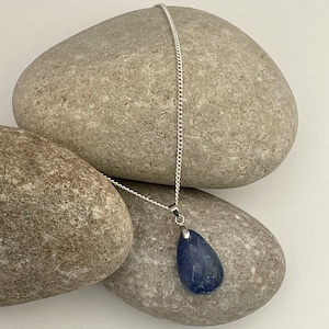 K&ouml;nnte beinhalten: Eine silberne Halskette mit einem tropfenf&ouml;rmigen, tiefblauen Edelsteinanh&auml;nger. Der Anh&auml;nger h&auml;ngt an einer feinen Silberkette. Die Halskette wird vor einem Hintergrund aus glatten, grauen Steinen pr&auml;sentiert.