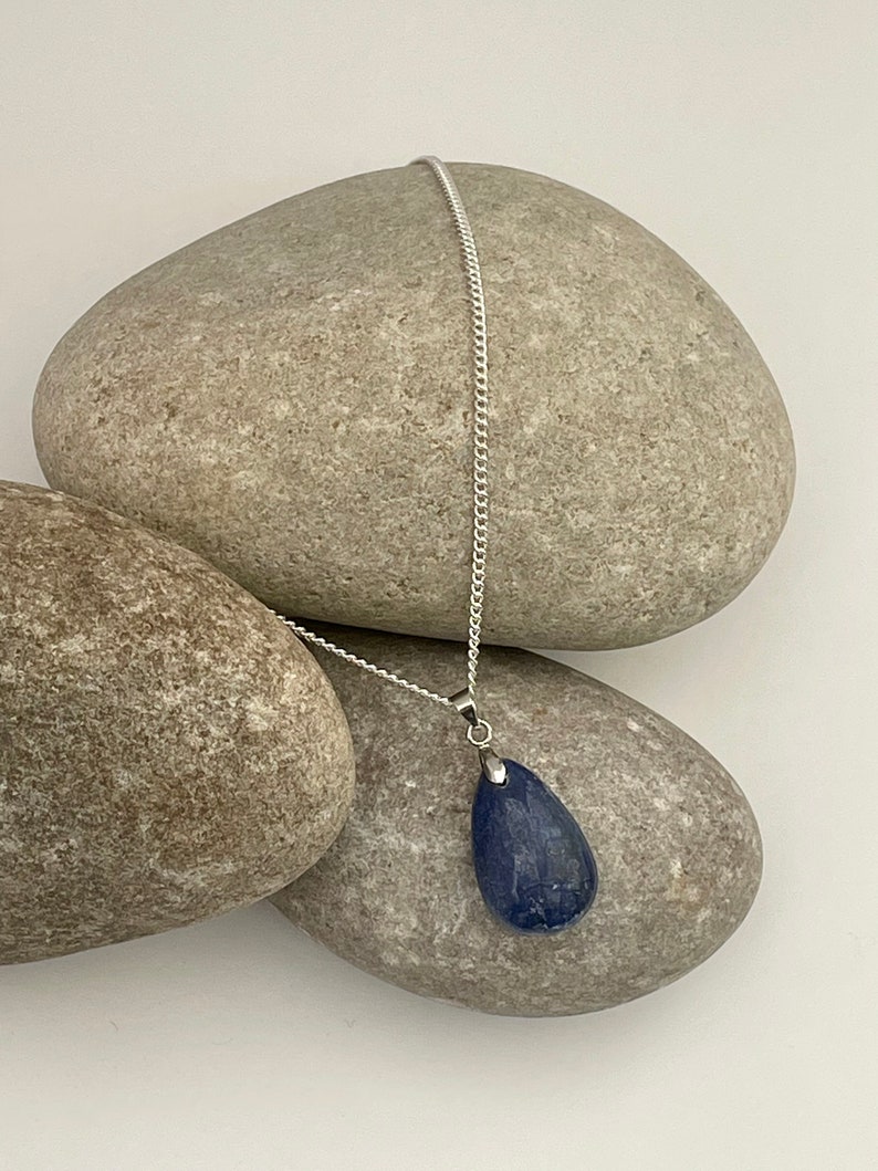 K&ouml;nnte beinhalten: Eine silberne Halskette mit einem tropfenf&ouml;rmigen, tiefblauen Edelsteinanh&auml;nger. Der Anh&auml;nger h&auml;ngt an einer feinen Silberkette. Die Halskette wird vor einem Hintergrund aus glatten, grauen Steinen pr&auml;sentiert.