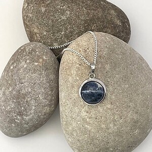 May include: A silver chain necklace with a round pendant featuring a blue and white stone. The necklace is displayed on top of three gray stones.