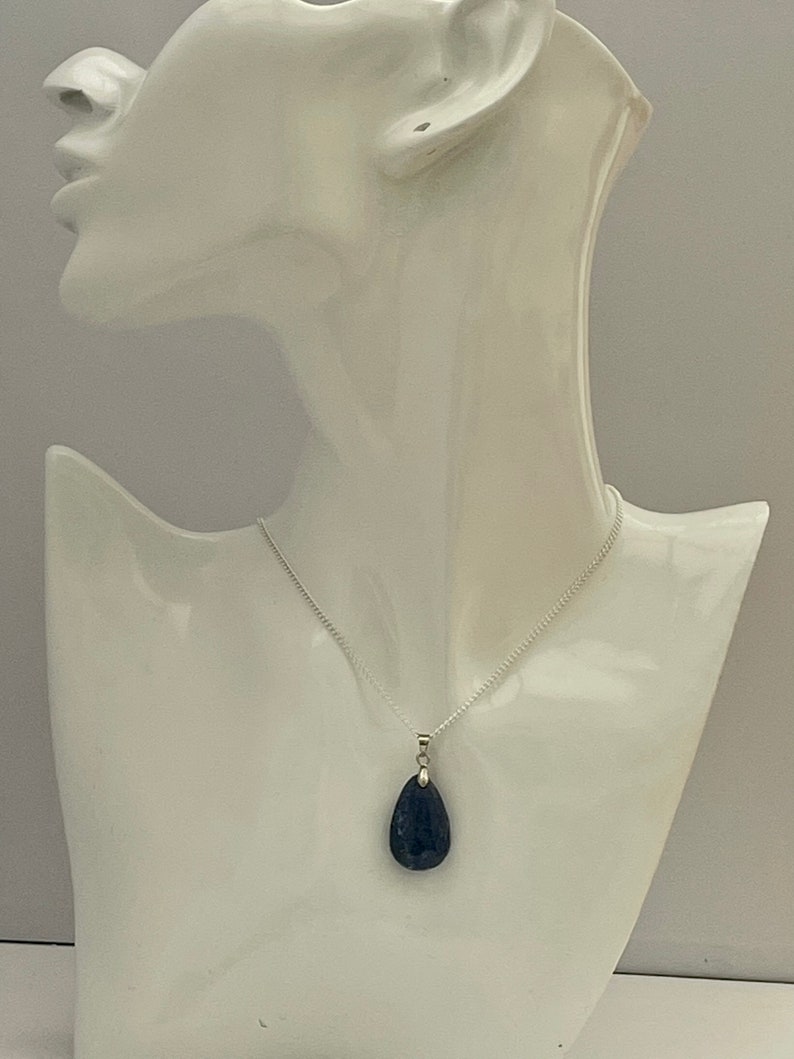K&ouml;nnte beinhalten: Eine silberne Halskette mit einem dunkelblauen, tropfenf&ouml;rmigen Anh&auml;nger. Der Anh&auml;nger h&auml;ngt an einer filigranen Kette und wird auf einer wei&szlig;en Mannequin-B&uuml;ste pr&auml;sentiert. Der Anh&auml;nger hat eine glatte, polierte Oberfl&auml;che.