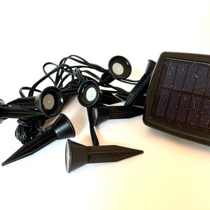 May include: Black solar-powered landscape spotlights with a solar panel. The spotlights have a conical shape and are connected by black wires. Each spotlight has a bright LED light. The solar panel is rectangular.