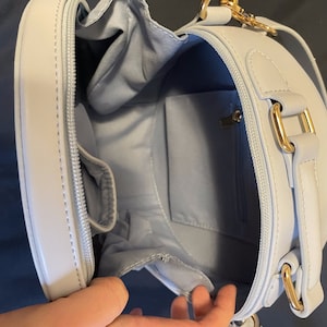 K&ouml;nnte beinhalten: Eine hellblaue runde Umh&auml;ngetasche mit einem goldenen Rei&szlig;verschluss und Beschl&auml;gen. Die Tasche hat eine kleine Innentasche.