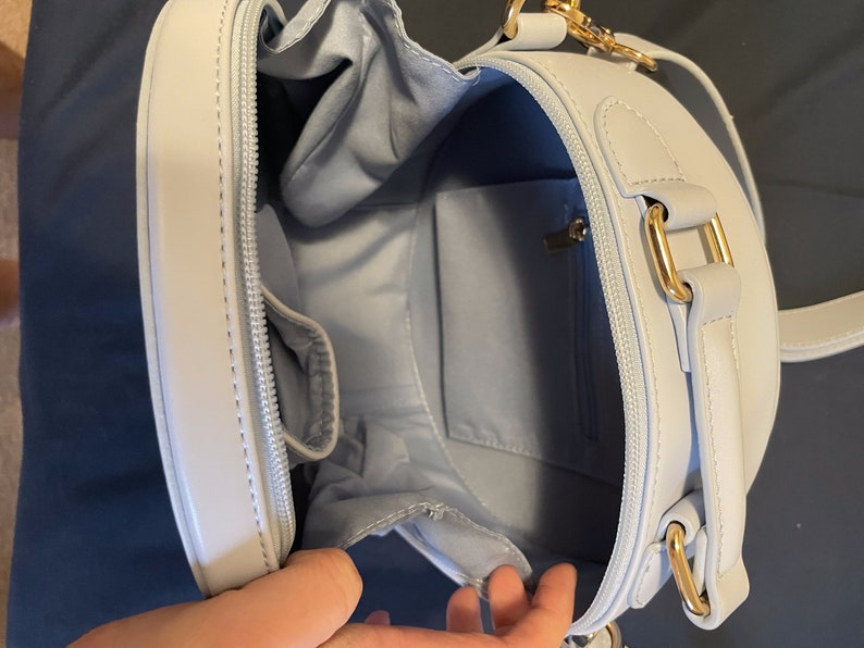 K&ouml;nnte beinhalten: Eine hellblaue runde Umh&auml;ngetasche mit einem goldenen Rei&szlig;verschluss und Beschl&auml;gen. Die Tasche hat eine kleine Innentasche.