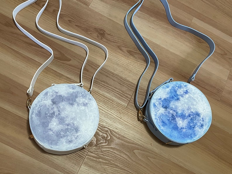 K&ouml;nnte beinhalten: Zwei runde mondf&ouml;rmige Handtaschen mit verstellbaren Tr&auml;gern. Eine Tasche ist wei&szlig; mit einem hellblauen Monddesign und die andere ist hellblau mit einem dunkelblauen Monddesign.
