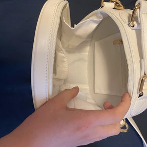 K&ouml;nnte beinhalten: Wei&szlig;e runde Handtasche mit goldenem Rei&szlig;verschluss und goldenem Kettenriemen. Die Tasche ist offen und das Innere ist mit wei&szlig;em Stoff ausgekleidet.