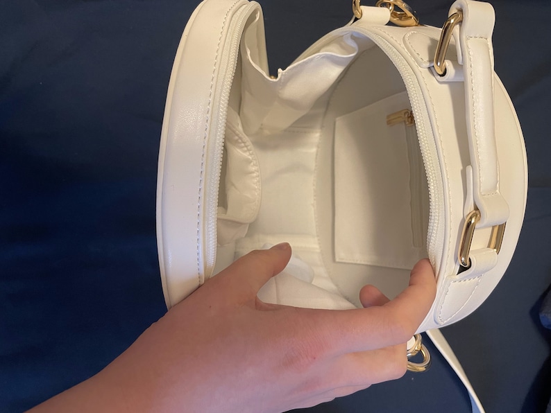 K&ouml;nnte beinhalten: Wei&szlig;e runde Handtasche mit goldenem Rei&szlig;verschluss und goldenem Kettenriemen. Die Tasche ist offen und das Innere ist mit wei&szlig;em Stoff ausgekleidet.