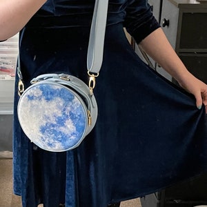 K&ouml;nnte beinhalten: Eine hellblaue runde Umh&auml;ngetasche mit einem wei&szlig;en und blauen Wolkenmuster. Die Tasche hat einen goldenen Kettenriemen und einen goldenen Verschluss.