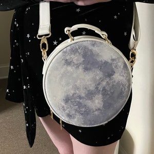 K&ouml;nnte beinhalten: Eine wei&szlig;e runde Handtasche mit einem Mond-Druckdesign. Die Tasche hat einen goldenen Kettenriemen und einen goldenen Verschluss.