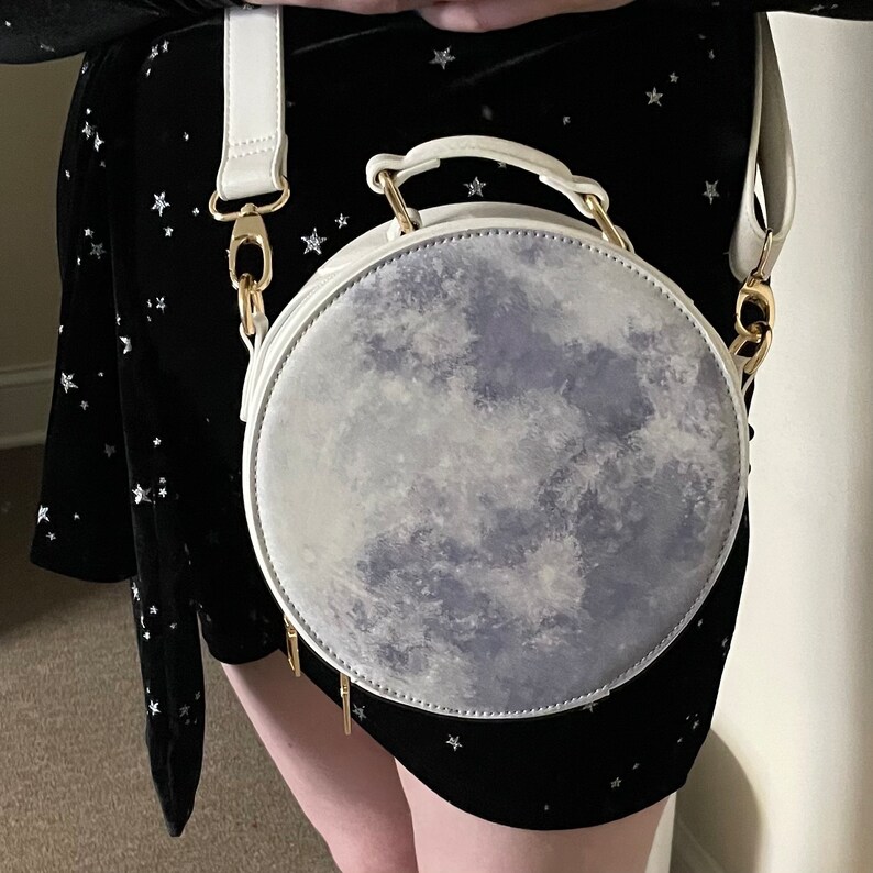 K&ouml;nnte beinhalten: Eine wei&szlig;e runde Handtasche mit einem Mond-Druckdesign. Die Tasche hat einen goldenen Kettenriemen und einen goldenen Verschluss.