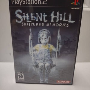 Puede incluir: Caja de juego de PlayStation 2 con un fondo negro y un título blanco que dice "Silent Hill Shattered Memories". La portada presenta a una niña sentada en un columpio cubierto de hielo. El juego está clasificado como M para Maduros por la ESRB y es publicado por Konami.