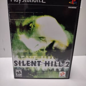 Puede incluir: Caja de juego de PlayStation 2 para Silent Hill 2. La caja es negra con una imagen verde y blanca de la cara de una mujer. El juego está clasificado como M para adultos por la PEGI.