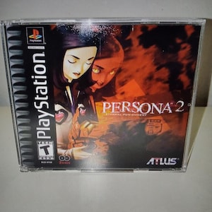 May include: A PlayStation game case for Persona 2: Eternal Punishment. The cover art features two female figures with dark hair and a fiery background. The PlayStation logo is visible on the left side, along with the Atlus logo.