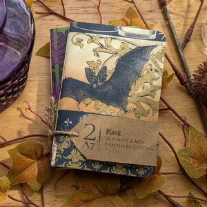 May include: Two A7 blank notebooks, pamphlet stitched, tied with twine. The covers feature a bat illustration with gold accents and a dark blue floral pattern. A purple glass bottle and decorative foliage are in the background.
