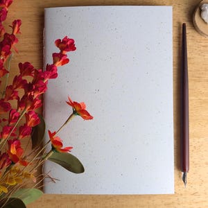 May include: A white notebook with a speckled texture, positioned next to a bouquet of vibrant red and orange flowers and a dark brown calligraphy pen with a metal nib. The notebook features a clean, blank cover and a stitched spine detail.